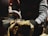 Hands wearing gloves hold a jar under a metal spout as a clear liquid pours into the jar. The background is blurred, focusing on the action in the foreground.