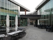 A modern architectural space with glass walls and angular roofs creates a sleek atmosphere. Several outdoor seating arrangements with white umbrellas and metal tables can be seen. The area appears to be part of a commercial or dining facility.