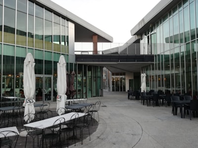 A modern architectural space with glass walls and angular roofs creates a sleek atmosphere. Several outdoor seating arrangements with white umbrellas and metal tables can be seen. The area appears to be part of a commercial or dining facility.