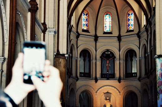An interior view of a church with arched architecture and stained glass windows at the top. A crucifix and a religious statue are visible at the front. Blurred hands are holding a smartphone in the foreground, capturing the scene.