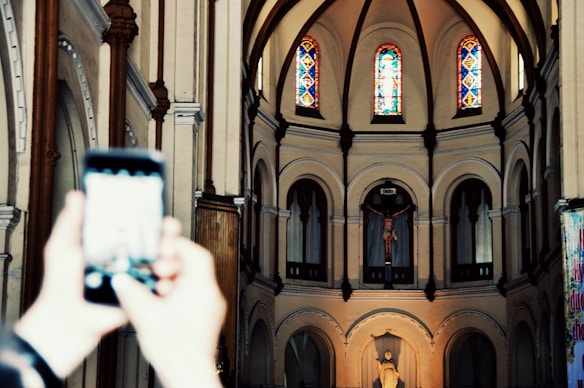 An interior view of a church with arched architecture and stained glass windows at the top. A crucifix and a religious statue are visible at the front. Blurred hands are holding a smartphone in the foreground, capturing the scene.