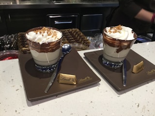 Two cups of dessert sit on plates with a small chocolate piece labeled 'Lindt' beside each. The dessert appears layered with whipped cream, chocolate sauce, and possibly nuts on top. Spoons are placed beside each cup, and the setting seems to be on a countertop with various containers in the background, suggesting a cafe or dessert shop.