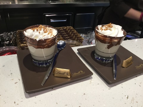 Two cups of dessert sit on plates with a small chocolate piece labeled 'Lindt' beside each. The dessert appears layered with whipped cream, chocolate sauce, and possibly nuts on top. Spoons are placed beside each cup, and the setting seems to be on a countertop with various containers in the background, suggesting a cafe or dessert shop.