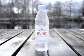 A clear plastic bottle of Evian natural mineral water standing upright on a wooden surface. The bottle is 500 ml in size and has water droplets on it. In the background, there is a blurred, wintry outdoor setting with bare trees and a body of water, adding a sense of cold and serenity to the image.