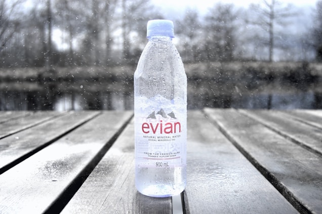 A clear plastic bottle of Evian natural mineral water standing upright on a wooden surface. The bottle is 500 ml in size and has water droplets on it. In the background, there is a blurred, wintry outdoor setting with bare trees and a body of water, adding a sense of cold and serenity to the image.