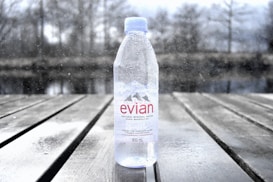 A clear plastic bottle of Evian natural mineral water standing upright on a wooden surface. The bottle is 500 ml in size and has water droplets on it. In the background, there is a blurred, wintry outdoor setting with bare trees and a body of water, adding a sense of cold and serenity to the image.