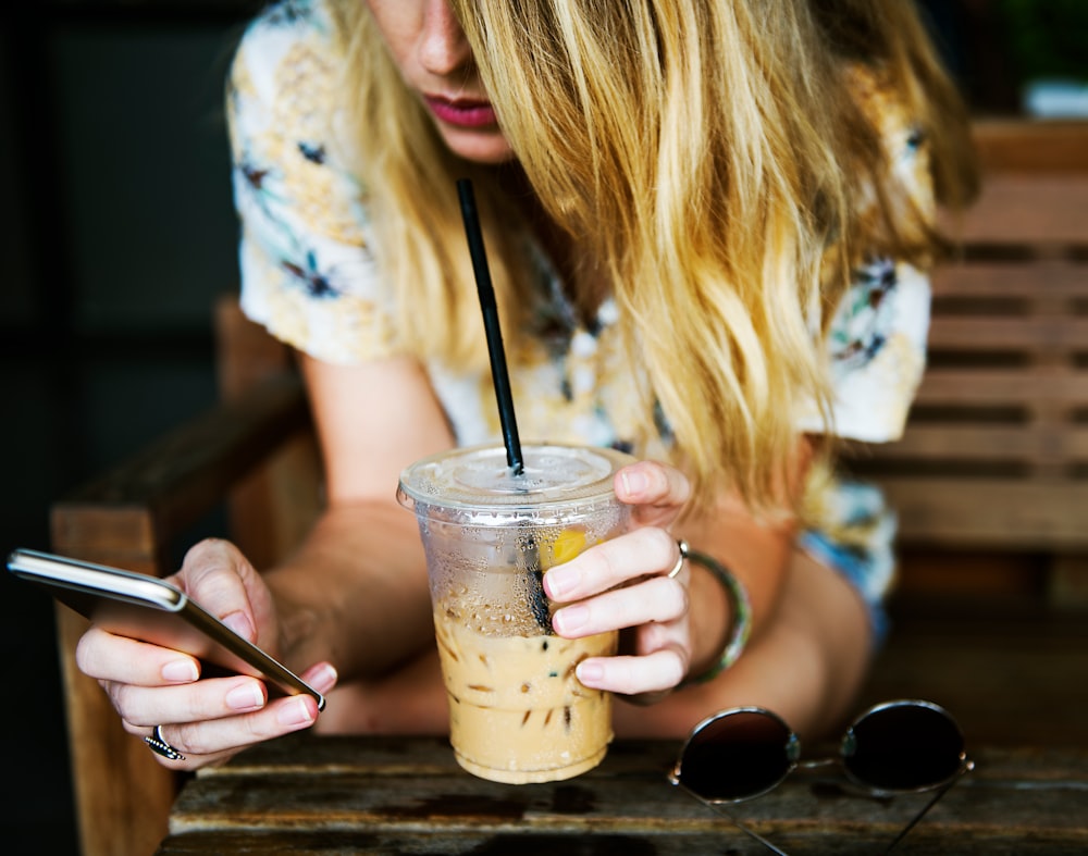 shallow focus photography of woman holding clear plastic disposable drinking cup while using smartphone
