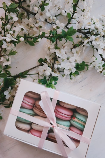 A white gift box filled with assorted macarons in pastel colors is tied with a pink ribbon. The box is surrounded by blossoming branches with delicate white flowers and green leaves, placed on a light wooden surface.