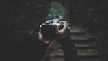 Hands holding a camera in a dimly lit forest setting with blurred steps in the background. The scene is captured in a dark, moody atmosphere.