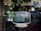 A vintage television with a static-filled screen sits on a small round table. It is displayed in a window surrounded by hanging green plants, with a reflection of a street scene in the glass.