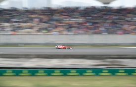 A red Formula 1 race car speeds along a racetrack with blurred spectators and seating in the background, indicating rapid motion and a dynamic event.