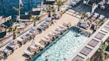 A luxurious poolside setting features numerous sun loungers and potted palm trees arranged on a wooden deck. Several people are enjoying the swimming pool, with some relaxing on the loungers. Adjacent to the pool area, there are boats docked by the water, under a clear blue sky, adding a nautical touch to the scene.