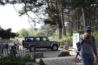 A rugged SUV is parked on a sandy path amid a forested area. The scene is surrounded by tall pine trees and leafy vegetation, creating a serene natural setting. Several people, including children, are gathered around, engaging in various outdoor activities. A person in the foreground appears to be pointing towards the camera while wearing casual attire.