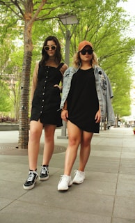Two young women stand confidently on a tree-lined sidewalk. They wear stylish outfits, including sunglasses, sneakers, and casual dresses. One has a denim jacket draped over her shoulders.