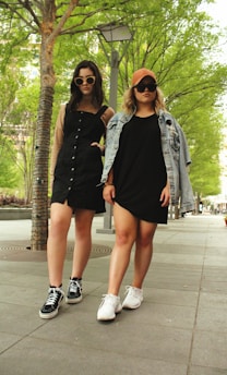 Two young women stand confidently on a tree-lined sidewalk. They wear stylish outfits, including sunglasses, sneakers, and casual dresses. One has a denim jacket draped over her shoulders.