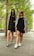 Two young women stand confidently on a tree-lined sidewalk. They wear stylish outfits, including sunglasses, sneakers, and casual dresses. One has a denim jacket draped over her shoulders.