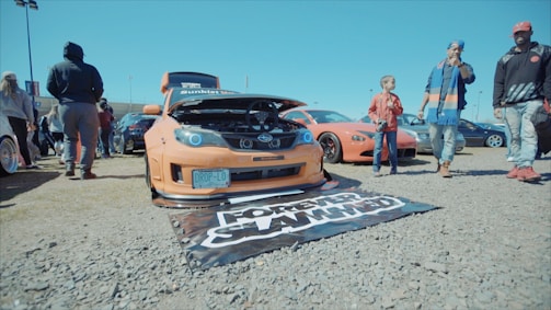 A modified orange sports car with its hood open is prominently displayed in a parking lot. Several people are gathered around, some observing the car while others are walking by. The car has a license plate reading 'DROP-LO' and a mat on the ground with a design. The scene is bright and appears to be a casual gathering or car show.