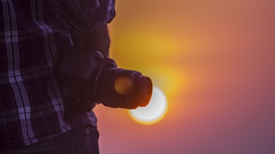 A photographer holds a camera against the backdrop of a vibrant sunset, with warm colors blending into the sky. The photographer is wearing a plaid shirt, and the scene exudes a sense of calm and creativity.
