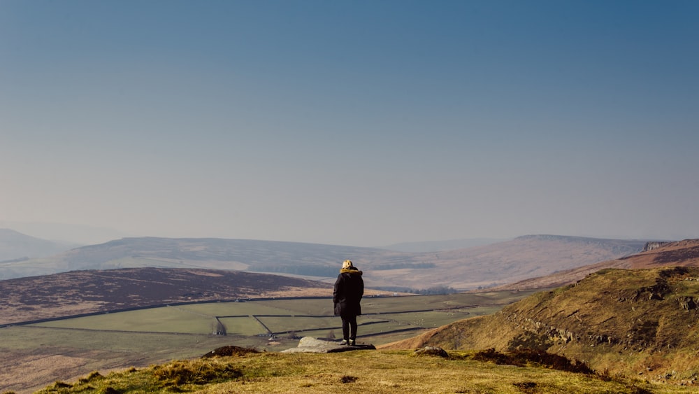 person wearing coat standing on cliff looking towards field under clear blue sky during daytime