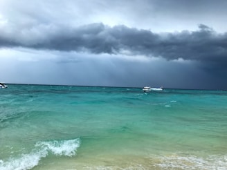 Dark storm clouds loom over a vast expanse of turquoise sea, creating a moody and dramatic atmosphere. The ocean waves gently lap against the shore, while a small boat is visible in the distance, appearing to navigate the changing weather conditions.