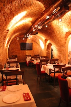 The image depicts an elegant restaurant interior with a vaulted brick ceiling and warm lighting. The tables are set with white tablecloths, red napkins, glassware, and cutlery, creating a cozy and inviting atmosphere. The seating consists of wooden chairs, and the room has an arched architecture feature, adding to the charm.