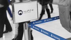 A sectioned off area indicated by blue PlayStation-branded tape. A display stand with the logo and text 'Meu PS4: Tudo sobre PS4 no Brasil' is visible. Several people are walking by, with one carrying a backpack on the right.