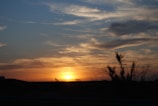 An expansive sky filled with dramatic clouds is illuminated by the warm hues of a sunset. Silhouetted against the horizon are two oil pump jacks, emphasizing an industrial landscape. Sparse vegetation is visible in the foreground, suggesting a desert or arid region.
