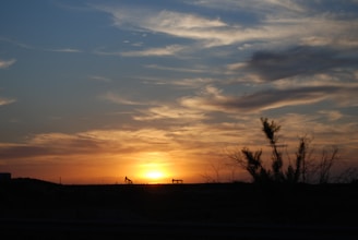 An expansive sky filled with dramatic clouds is illuminated by the warm hues of a sunset. Silhouetted against the horizon are two oil pump jacks, emphasizing an industrial landscape. Sparse vegetation is visible in the foreground, suggesting a desert or arid region.