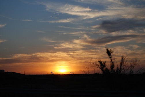 An expansive sky filled with dramatic clouds is illuminated by the warm hues of a sunset. Silhouetted against the horizon are two oil pump jacks, emphasizing an industrial landscape. Sparse vegetation is visible in the foreground, suggesting a desert or arid region.