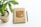 A spiral-bound notebook with a brown kraft cover lies on a white surface next to a black pen labeled 'envato'. The notebook features graphic text with a McDonald's theme and logos including 'McCafe', 'BicMac', and 'Saigon'. Beside the notebook, a small potted plant with green leaves is visible.