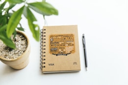 A spiral-bound notebook with a brown kraft cover lies on a white surface next to a black pen labeled 'envato'. The notebook features graphic text with a McDonald's theme and logos including 'McCafe', 'BicMac', and 'Saigon'. Beside the notebook, a small potted plant with green leaves is visible.
