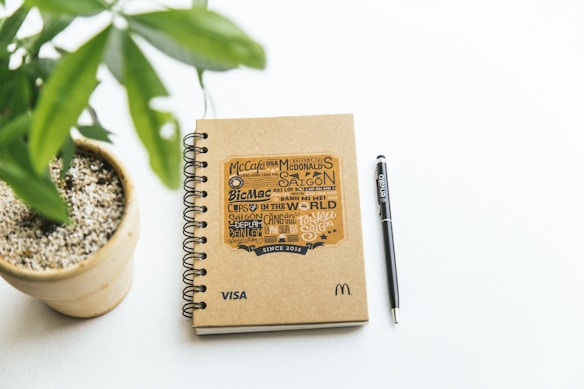 A spiral-bound notebook with a brown kraft cover lies on a white surface next to a black pen labeled 'envato'. The notebook features graphic text with a McDonald's theme and logos including 'McCafe', 'BicMac', and 'Saigon'. Beside the notebook, a small potted plant with green leaves is visible.