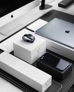 A sleek, modern workspace featuring a collection of neatly arranged Apple products. Items include an iMac with a keyboard, a MacBook Pro, an Apple Watch placed on its box, and an iPhone on a black box. The products are set on a black and white tabletop, reflecting a minimalist and organized aesthetic.