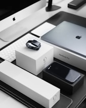 A sleek, modern workspace featuring a collection of neatly arranged Apple products. Items include an iMac with a keyboard, a MacBook Pro, an Apple Watch placed on its box, and an iPhone on a black box. The products are set on a black and white tabletop, reflecting a minimalist and organized aesthetic.
