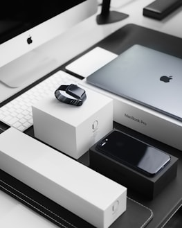 A sleek, modern workspace featuring a collection of neatly arranged Apple products. Items include an iMac with a keyboard, a MacBook Pro, an Apple Watch placed on its box, and an iPhone on a black box. The products are set on a black and white tabletop, reflecting a minimalist and organized aesthetic.