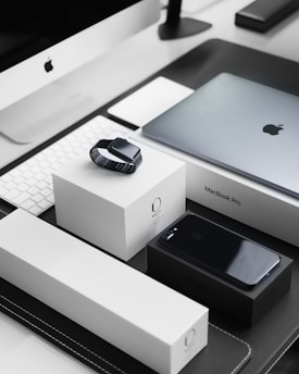A sleek, modern workspace featuring a collection of neatly arranged Apple products. Items include an iMac with a keyboard, a MacBook Pro, an Apple Watch placed on its box, and an iPhone on a black box. The products are set on a black and white tabletop, reflecting a minimalist and organized aesthetic.