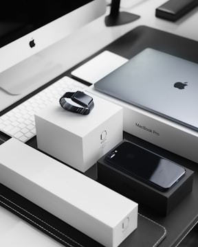 A sleek, modern workspace featuring a collection of neatly arranged Apple products. Items include an iMac with a keyboard, a MacBook Pro, an Apple Watch placed on its box, and an iPhone on a black box. The products are set on a black and white tabletop, reflecting a minimalist and organized aesthetic.