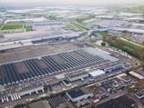 Aerial view of a large industrial complex featuring extensive warehouses with flat and pitched roofs. Numerous trucks and vehicles are parked around the complex, indicating logistics or manufacturing activities. Surrounding the industrial area are green fields and smaller buildings, with roads connecting different sections of the site. The atmosphere appears clear, with farmlands visible in the distance.