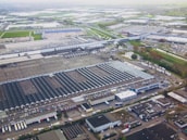 Aerial view of a large industrial complex featuring extensive warehouses with flat and pitched roofs. Numerous trucks and vehicles are parked around the complex, indicating logistics or manufacturing activities. Surrounding the industrial area are green fields and smaller buildings, with roads connecting different sections of the site. The atmosphere appears clear, with farmlands visible in the distance.