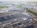 Aerial view of a large industrial complex featuring extensive warehouses with flat and pitched roofs. Numerous trucks and vehicles are parked around the complex, indicating logistics or manufacturing activities. Surrounding the industrial area are green fields and smaller buildings, with roads connecting different sections of the site. The atmosphere appears clear, with farmlands visible in the distance.