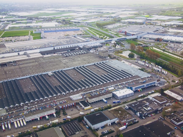 Aerial view of a large industrial complex featuring extensive warehouses with flat and pitched roofs. Numerous trucks and vehicles are parked around the complex, indicating logistics or manufacturing activities. Surrounding the industrial area are green fields and smaller buildings, with roads connecting different sections of the site. The atmosphere appears clear, with farmlands visible in the distance.
