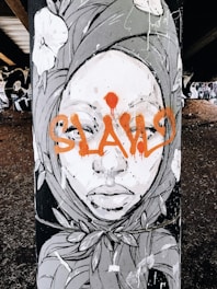 A piece of street art featuring a stylized face of a person adorned with a headscarf. The artwork includes floral elements and bold orange graffiti text across the face.