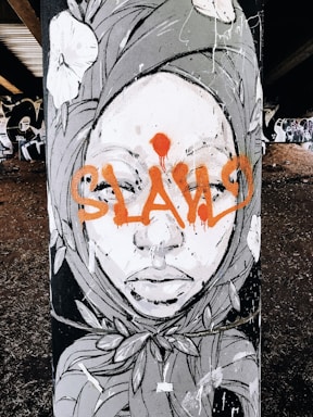 A piece of street art featuring a stylized face of a person adorned with a headscarf. The artwork includes floral elements and bold orange graffiti text across the face.