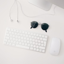 A clean white desk with a keyboard, earphones and sunglasses creating a calm, clear space