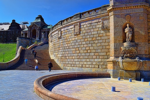 A historic architectural site featuring a large, ornate stone terrace with a statue embedded in the wall. Several people are walking along the wide stone steps and open plaza area. The building in the background has a classical style, and the area is bathed in sunlight with clear blue skies.
