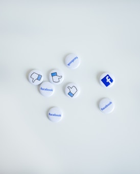 Several small round buttons are scattered on a white surface. The buttons feature various symbols and words associated with Facebook, including thumbs up, thumbs down, and the Facebook logo.