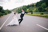 A couple dressed in wedding attire playfully interact on an empty road. The groom jumps enthusiastically while holding a bouquet of yellow flowers, and the bride runs beside him, smiling. The backdrop features lush green hills and trees, with a dramatic sky overhead.