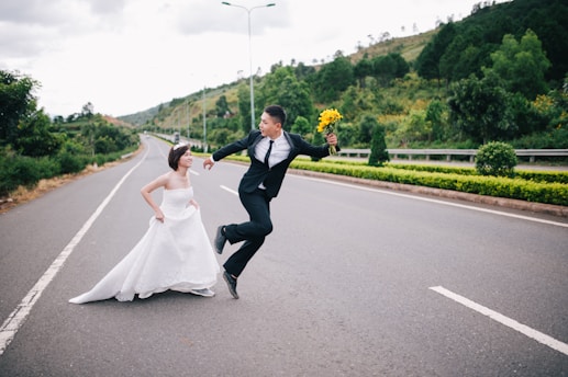 A couple dressed in wedding attire playfully interact on an empty road. The groom jumps enthusiastically while holding a bouquet of yellow flowers, and the bride runs beside him, smiling. The backdrop features lush green hills and trees, with a dramatic sky overhead.