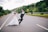 A couple dressed in wedding attire playfully interact on an empty road. The groom jumps enthusiastically while holding a bouquet of yellow flowers, and the bride runs beside him, smiling. The backdrop features lush green hills and trees, with a dramatic sky overhead.