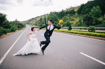A couple dressed in wedding attire playfully interact on an empty road. The groom jumps enthusiastically while holding a bouquet of yellow flowers, and the bride runs beside him, smiling. The backdrop features lush green hills and trees, with a dramatic sky overhead.